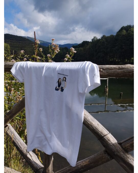 T-shirt bianca iaiadesign drappeggiata su una staccionata in legno con sfondo lago e fiori di campo in primo piano