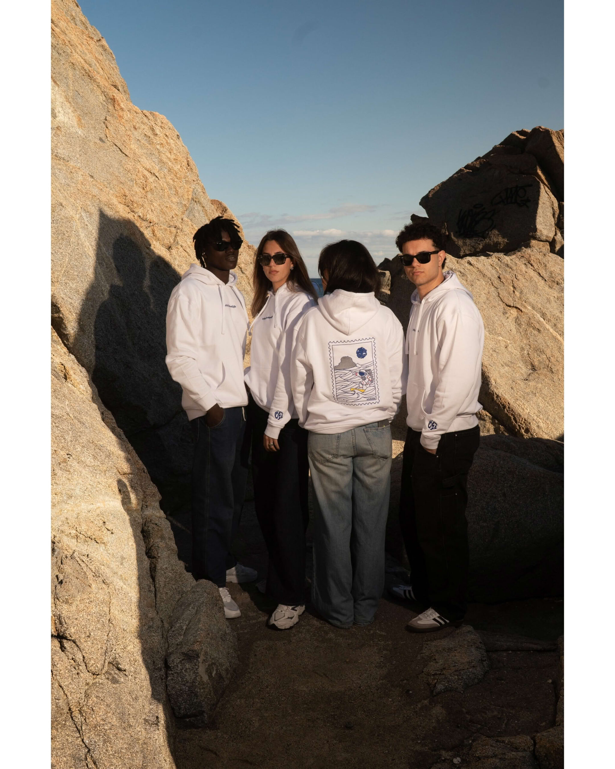 Gruppo di quattro ragazzi in piedi tra gli scogli al tramonto indossano felpa bianca con ricami blu.