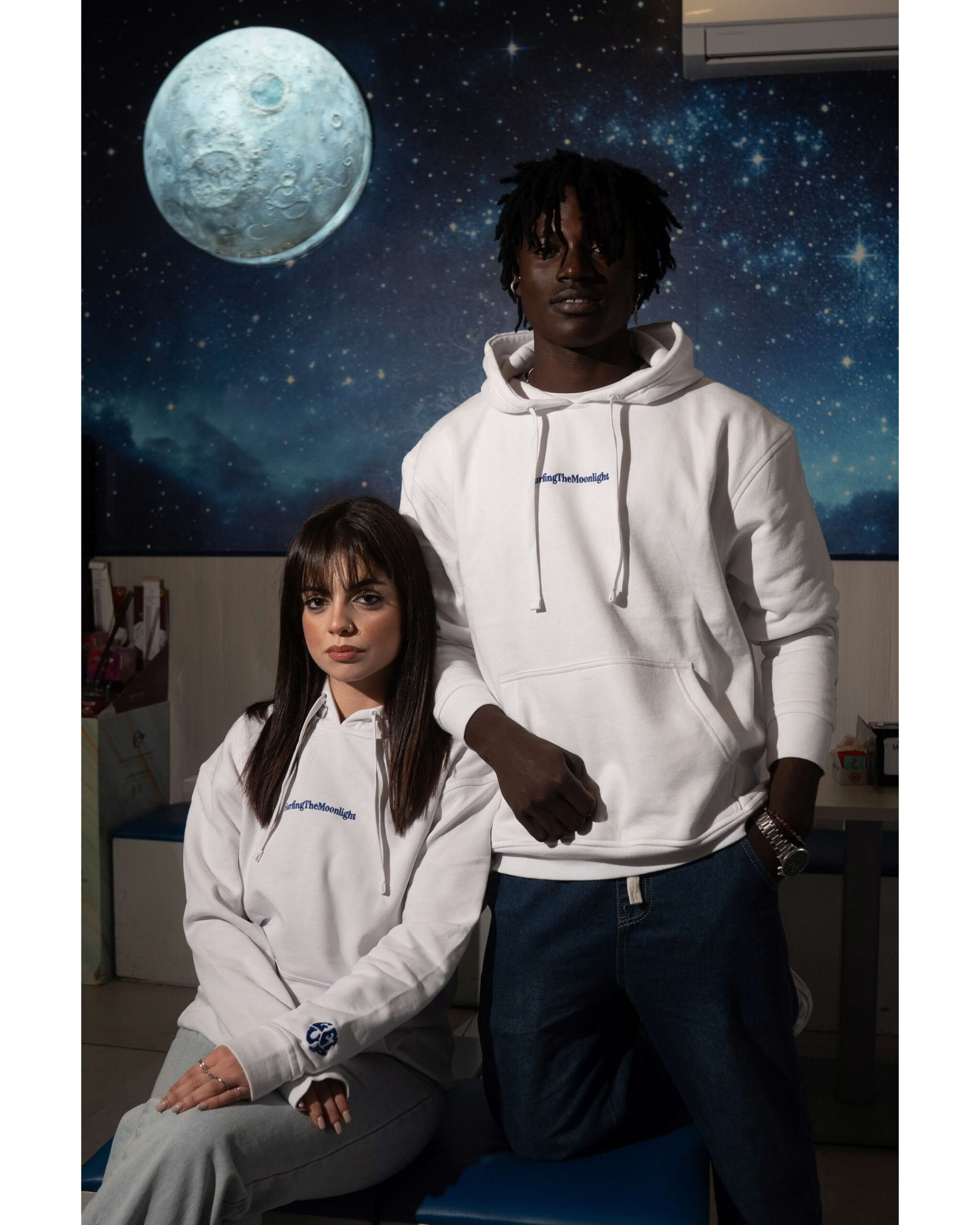 Ragazzo e ragazza in posa studio con sfondo luna gigante e stelle indossano felpa bianca con ricamo blu
