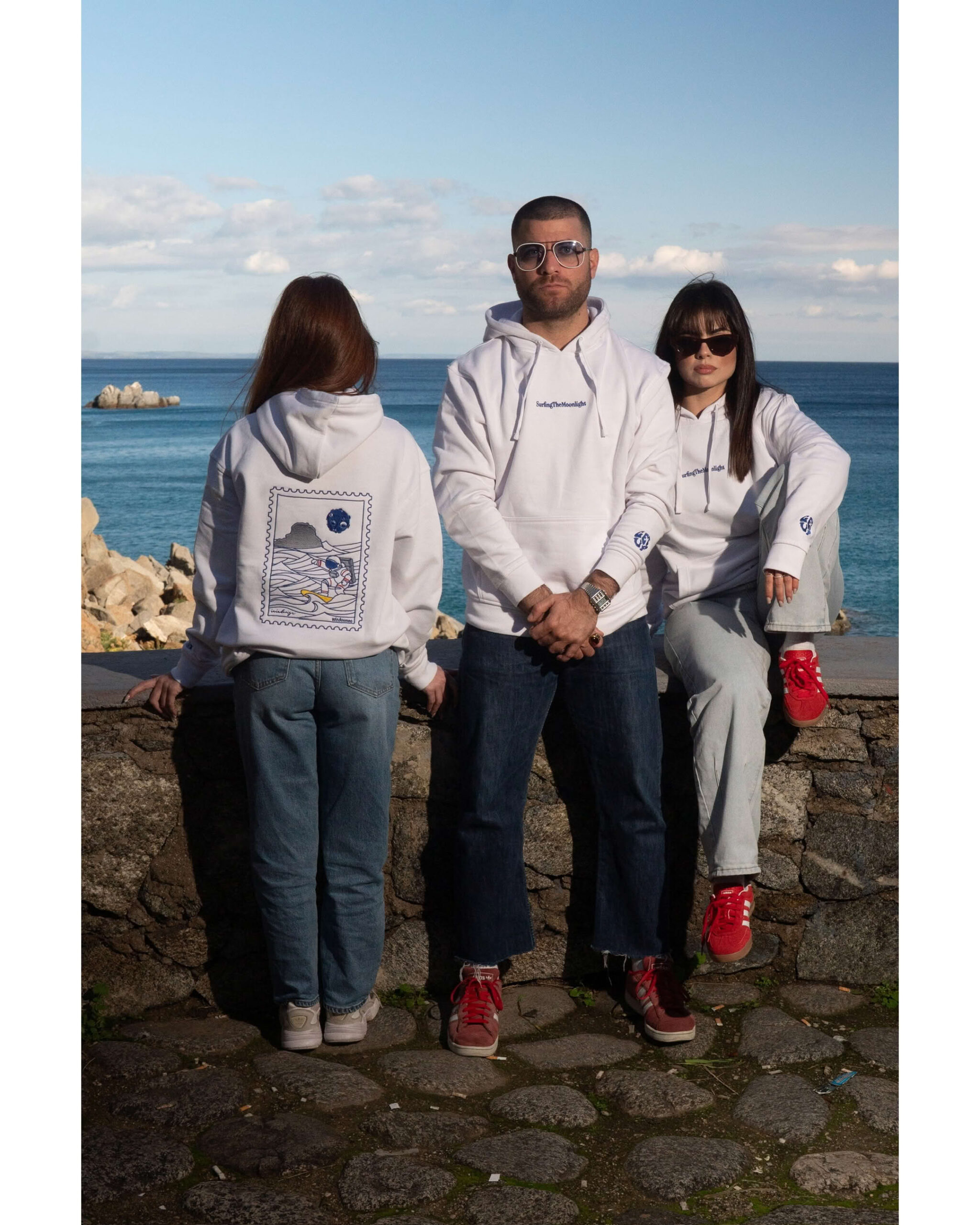 Gruppo di tre amici su muretto lungomare con felpe bianche coordinate, vista fronte e retro.