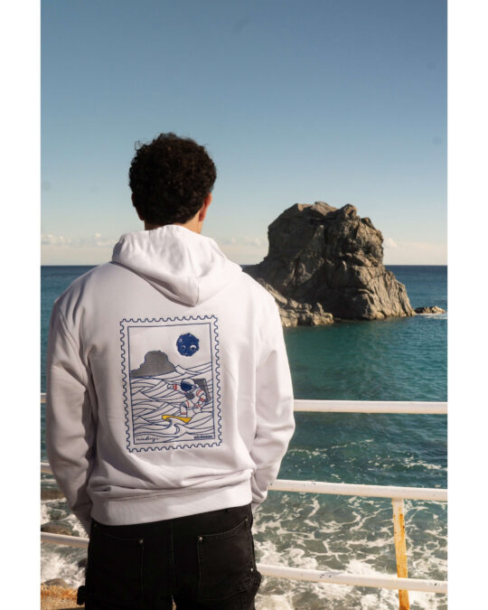 Ragazzo di spalle fronte mare guarda scoglio e indossa felpa bianca con ricamo posteriore astronauta surfista.