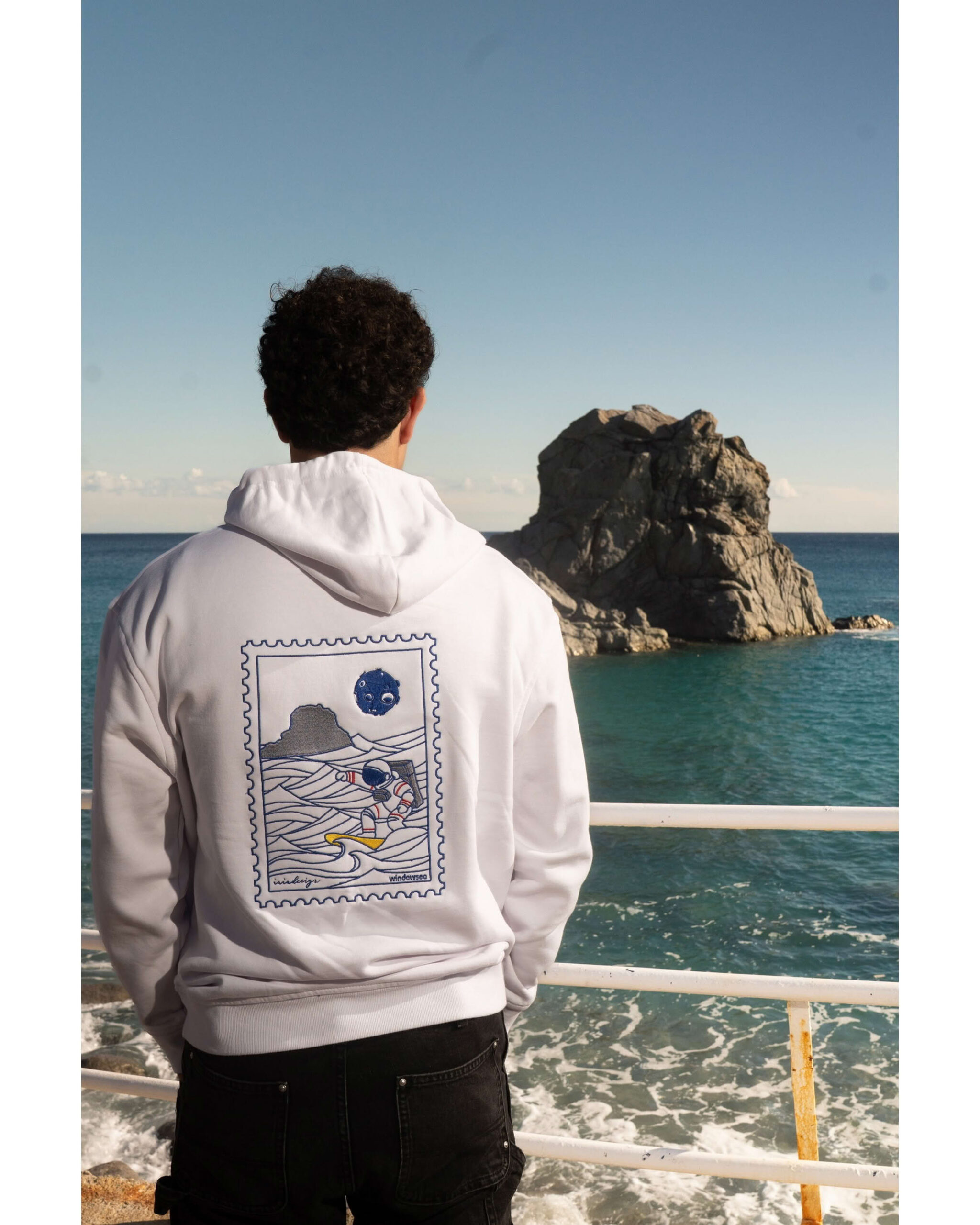 Ragazzo di spalle fronte mare guarda scoglio e indossa felpa bianca con ricamo posteriore astronauta surfista.