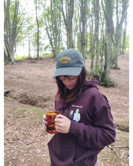 Donna nel bosco indossa felpa bordeaux con cappello Harvard e guarda in basso tenendo una tazza ambrata