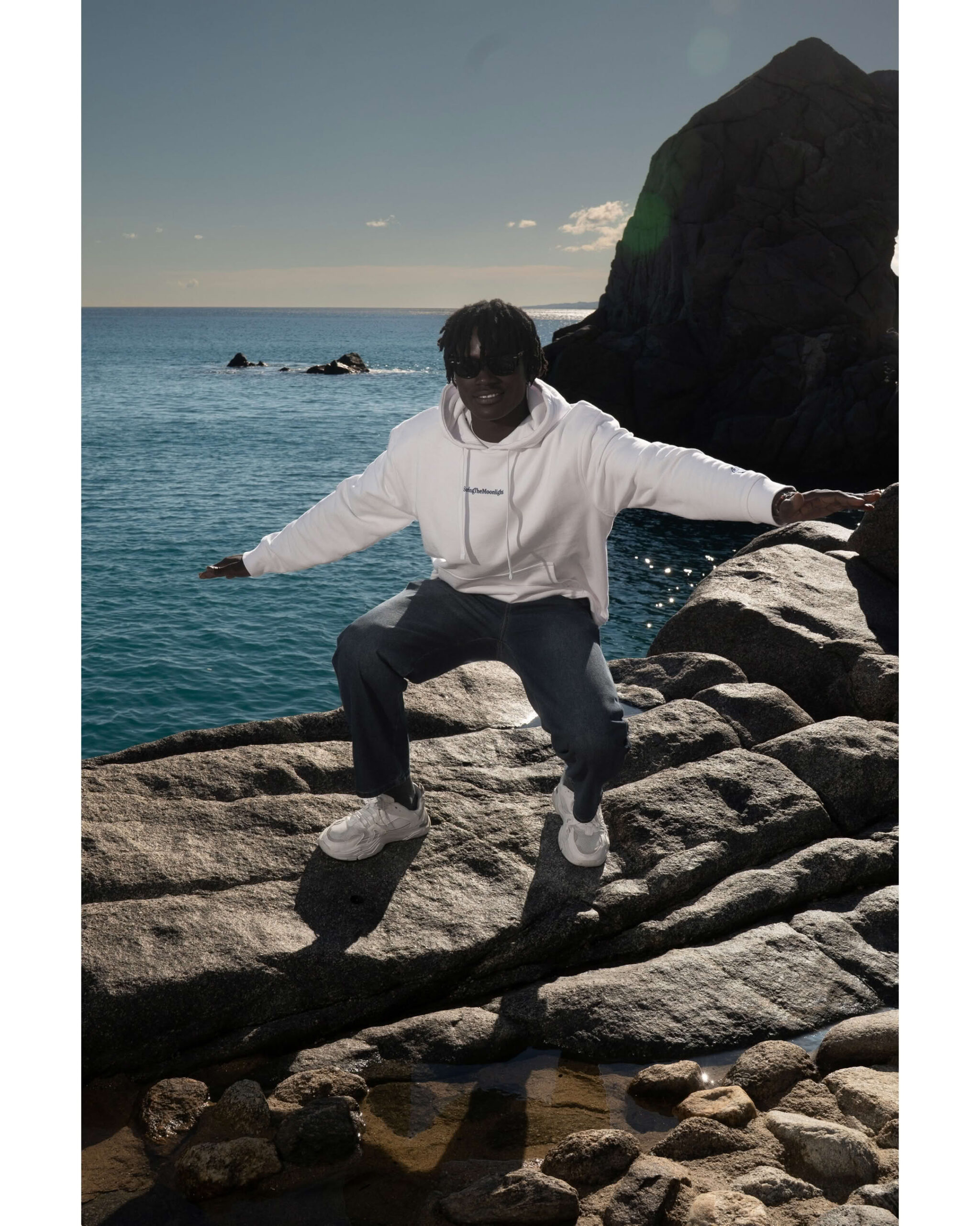 Ragazzo in posa dinamica su scoglio fronte mare indossa felpa bianca con scritta blu SurfingTheMoonlight.