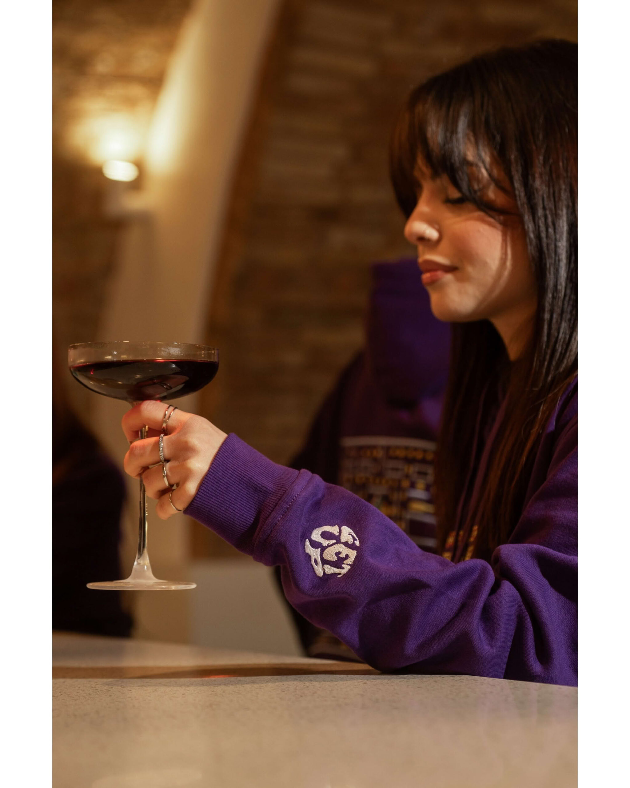 Ragazza di profilo beve un cocktail indossando felpa viola, focus sul ricamo bianco Get Up sulla manica.