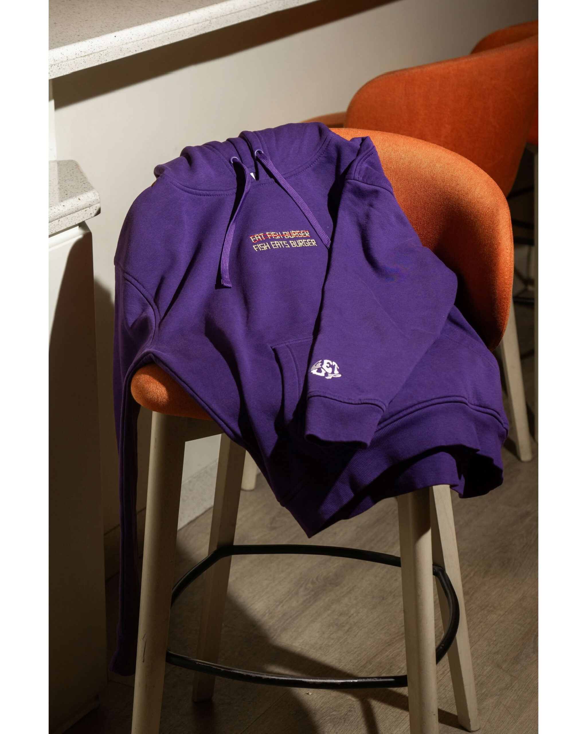 Felpa viola drappeggiata su sgabello arancione, visibile il ricamo frontale e il dettaglio manica Get Up.