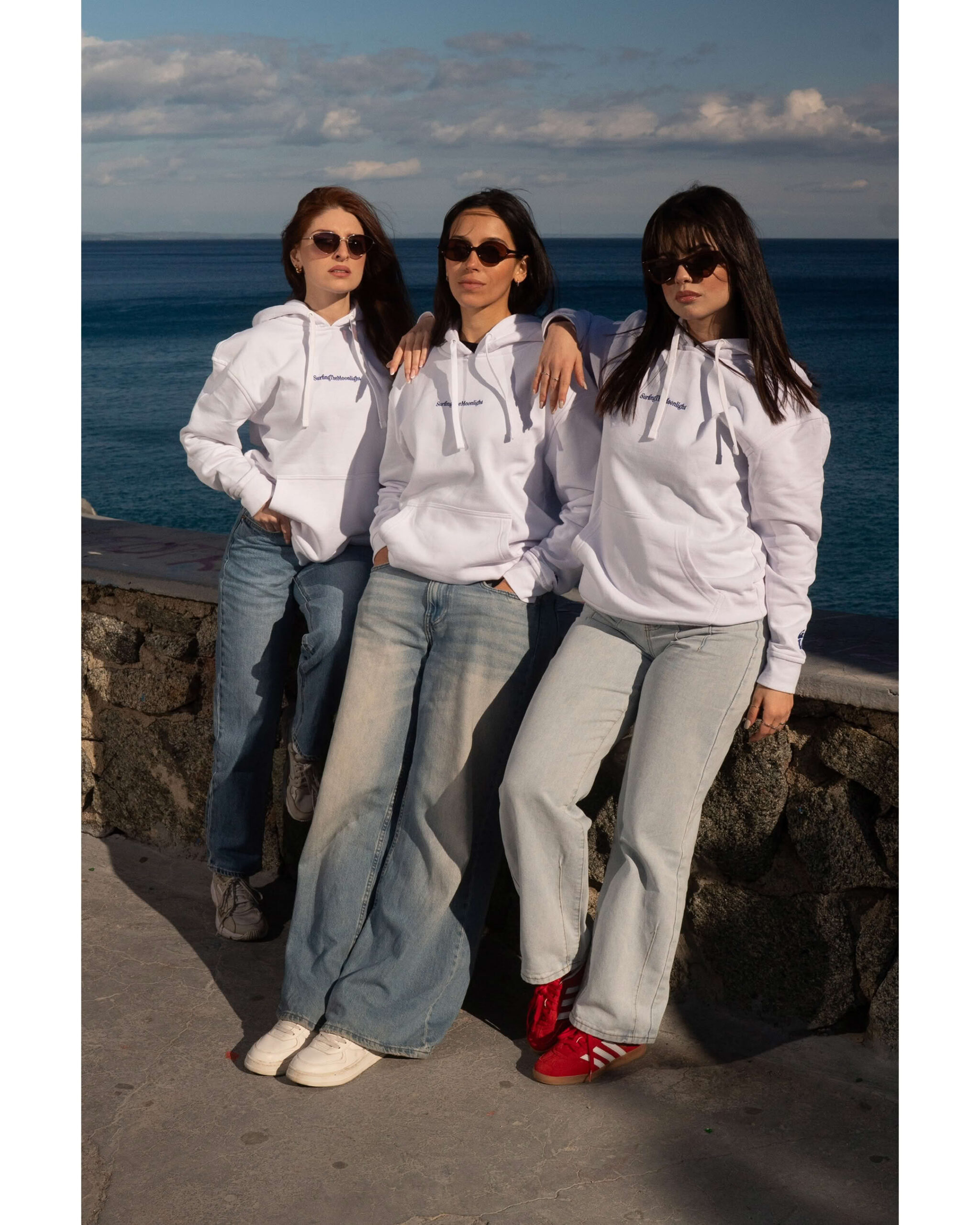 Tre ragazze in posa fronte mare indossano felpe bianche SurfingTheMoonlight e occhiali da sole