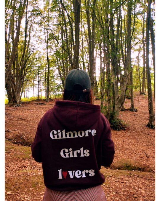 Vista posteriore di donna nel bosco con felpa bordeaux e grande scritta bianca Gilmore Girls lovers