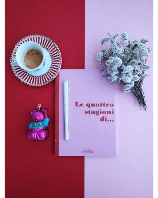 Planner rosa al centro su fondo bicolore rosso/rosa, con caffè, ornamento orso fucsia e fiori bianchi